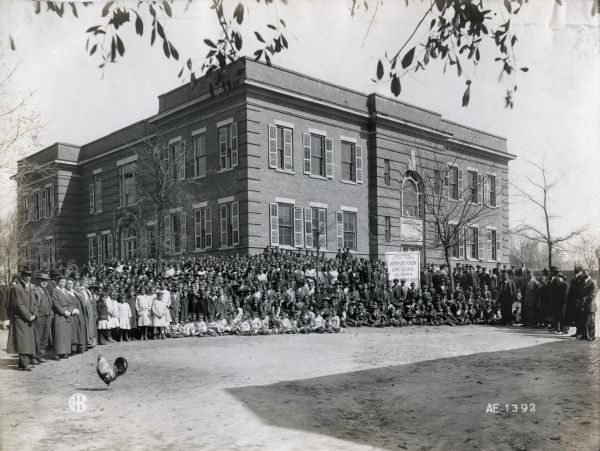 A large group of African American children stand with a sign reading: "Diversification of Livestock in Alabama" in front of the Montgomery State Normal School. Adults stand on either side of the children and a rooster stands on the drive in the left foreground.