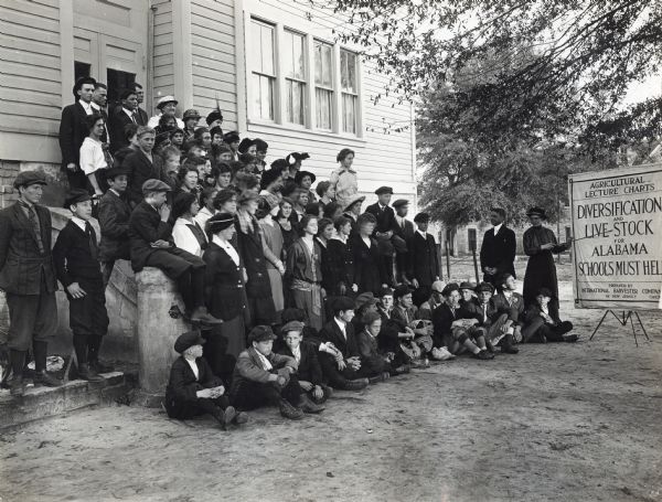 A group of children and adults gathered on the steps at Western Alabama Agricultural School to listen as Miss Zella Wigent presents on the subject of livestock diversification. She uses a pointer to direct the audience's attention toward a visual aid which reads: "Agricultural Lecture Charts. Diversification and Live Stock for Alabama. Schools Must Help. Prepared by International Harvester Company of New Jersey. Chicago."