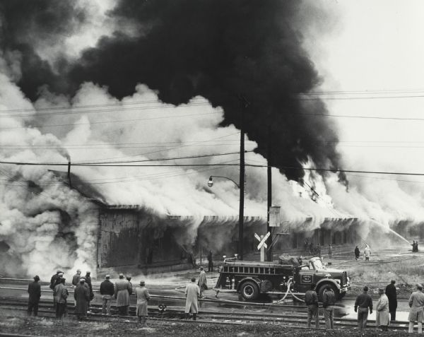 Men and fire fighters are gathered around burning building attempting to extinguish flames with an International fire truck. The truck is labeled "Engine No. 1 Company."