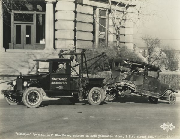 A man is towing a damaged automobile with an International Six-Speed Special truck. Original caption reads: "'Six speed special, 124" wheelbase, mounted on 30x5 pneumatic tires, IHC closed cab."