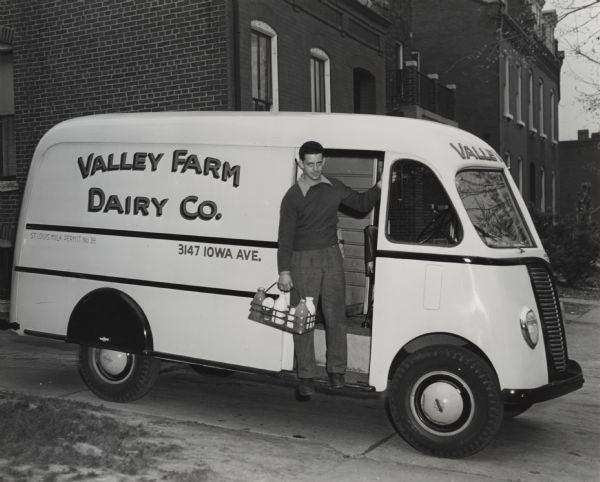 International truck with a metro body used by Valley Farm Dairy Company. A delivery man is standing on the foot board of the truck carrying a basket of bottles of milk.