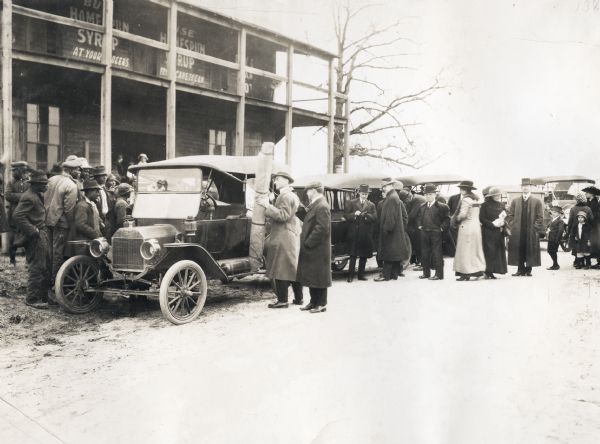 A group of men, women, and children leaving an agricultural presentation by J.E. Hite at Haile's store. Hite, dressed in a hat and overcoat, is loading a rolled chart into an automobile while groups of people are standing nearby. The signs hanging from the upper story of the building read: "Buy Homespun Syrup at Your Grocers" and "Use Homespun Syrup From Cane to Can."