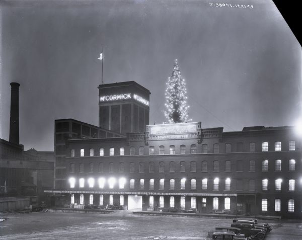 Elevated view of International Harvester's McCormick Works lit up on a December night. A very large lit Christmas tree is on the roof of the building. A sign along the front of the building reads: "'Quality is the foundation of our business!' — Alex Legge, President." The McCormick Works was built in 1873 and was located at Blue Island and Western Avenues in the Chicago subdivision called "Canalport." The factory closed in 1961. The original caption said: "Superintendent F.H. Harrison of McCormick works and his organization had their own Christmas tree. It was large enough to be visible to the employees for a considerable distance in all directions as they came to and went from their work. It was lighted and decorated in attractive style and exemplified the get-together spirit that is so characteristic of McCormick."