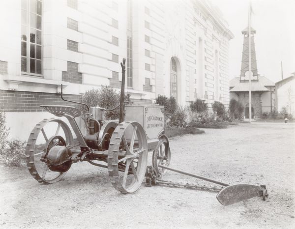 Three-quarter rear view of a McCormick Auto-Mower near a building at the Paris Exposition in France. The Auto-Mower was an experimental engine-powered mower that won first prize at the exposition. This is one of two Auto-Mowers produced by the company. This machine had a single-cylinder engine. The other machine had a two-cylinder engine.