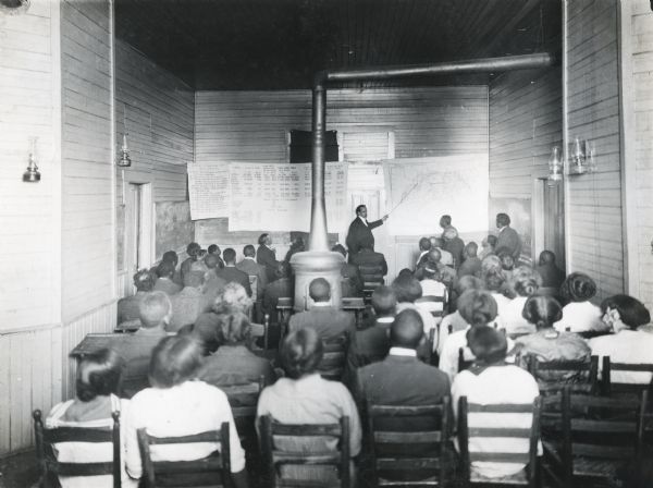 View from back of classroom of Dr. W.B. Hinds pointing at a graph as he delivers an agricultural lecture on cotton to students seated at North Alabama Baptist Academy. Additional charts and graphs line the front wall and a potbellied stove stands in the middle of the classroom.
