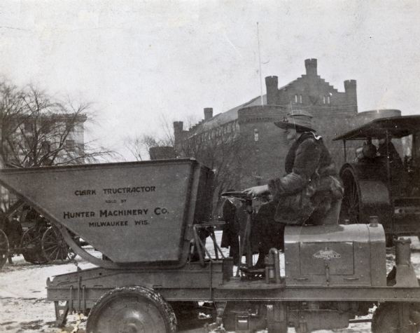 A young woman, most likely Esther Van Wagoner (Tufty), sitting on a Clark TrucTractor on the Library Mall of the University of Wisconsin-Madison campus. The tractor was sold by Hunter Machinery Company of Milwaukee. Other tractors, as well as the Red Gym or Armory campus building, are in the background.