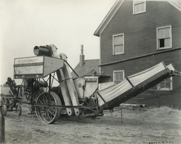 A McCormick-Deering Harvester No. 8 harvester-thresher (combine) parked outside a farmhouse. Decals and/or stencils are on the machine.
