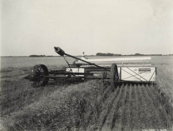 Rear view of man driving a tractor and pulling a McCormick-Deering windrow harvester in field. Decals are on the back of the machine.