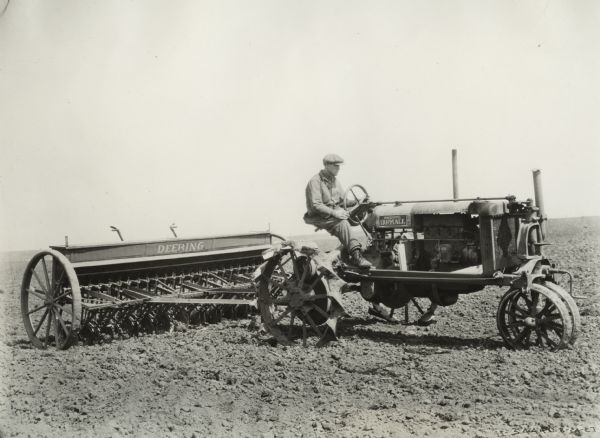 Side view of man driving Farmall Regular tractor pulling a Deering grain drill. Decals and/or stencils are on the tractor and grain drill.