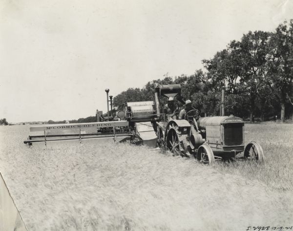 Three-quarter view from front of left side of a McCormick-Deering harvester-thresher (combine) being driven through a field by a man on a McCormick-Deering tractor. Decals and/or stencils are on both the tractor and harvester-thresher.