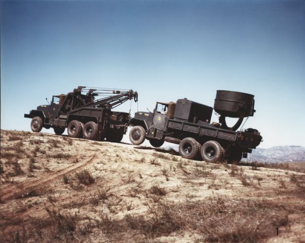 Color photograph of an M-40 Marine Corps vehicle equipped with a wrecker body towing a searchlight truck up a sandy hill. Mountains are in the far background. The specifications of the M-40 are as follows: 179" WB Truck, Chassis, with 11.00 x 20 front & dual rear tires."