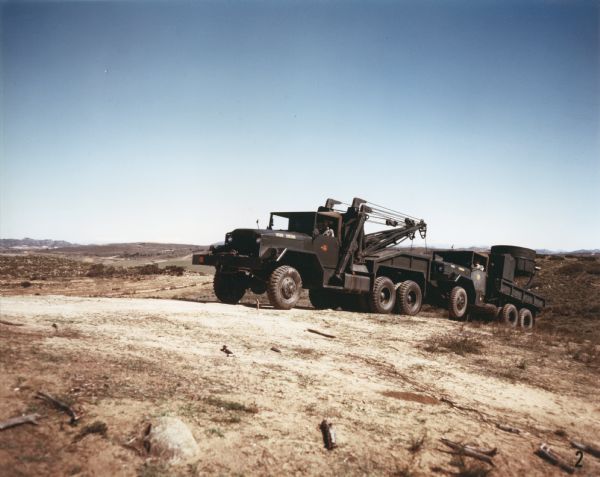 Color photograph of an M-40 Marine Corps vehicle equipped with a wrecker body towing a searchlight truck up a hill in an area of brush. Mountains and pastureland are in the far background. The specifications of the M-40 are as follows: 179" WB Truck, Chassis, with 11.00 x 20 front & dual rear tires."