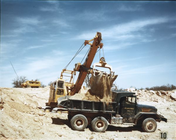 Color photograph of a model M-51 dump truck depositing raw lime rock from a quarry into the back of a truck marked "U.S. Air Force." Another vehicle is in the background. The specifications of the M-51 are as follows: 167" WB Truck, Dump, with 11.00 x 20 front & dual rear tires.