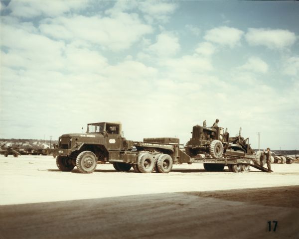 Color photograph of men loading a piece of construction equipment onto a trailer attached to a Model M-52 truck-tractor. Other vehicles parked on a gravel lot are in the background. The specifications of the M-52 are as follows: 167" WB Truck, Tractor, with 11.00 x 20 front and dual rear tires.
