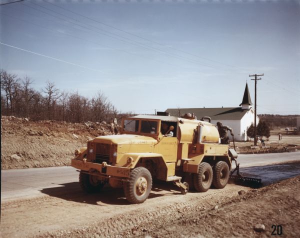 View towards of men using an M-61 to spread asphalt at Wolters Air Force Base. A building, possibly a church or chapel, stands in the background along the opposite side of the road. The specifications of the M-61 are as follows: 167" WB Truck, Chassis, with 11.00 x 20 front & dual rear tires.