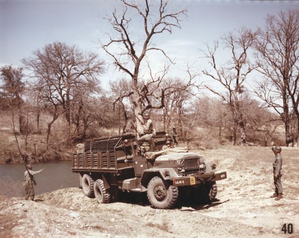 Color photograph of an M-139 truck transporting members of a bridge-building unit and construction materials to the bank of a river in a wooded area. The specifications of the M-139 are as follows: 215" WB Truck, Chassis, with 14.00 x 20 front & dual rear tires.