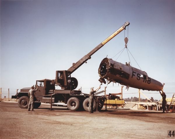 Color photograph of men guiding a piece of wreckage from a jet fighter onto the ground as it is lifted by the arm of a Model M-246 truck. The specifications of the M-246 are as follows: 215" WB Truck, Tractor Wrecker, with 12.00 x 20 front & dual rear tires.