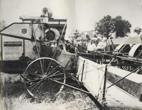 Side view of a McCormick-Deering harvester-thresher (combine). A group of men are standing behind the machine; and decals and/or stencils are on the side of the combine.