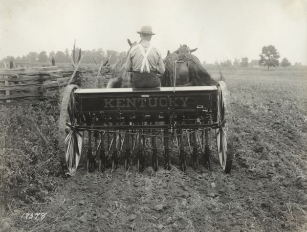 Rear view of man operating a Kentucky grain drill with a team of two horses. A decal with the "Kentucky" name is on the back of the drill.
