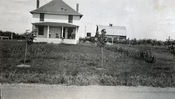 View from road of several people standing on the porch of an IH  demonstration farmhouse. A barn is in the background on the right, with a painted sign on its side reading: "Demonstration Farm."