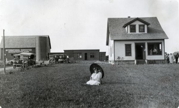 View of a woman posing holding a parasol while sitting on the front lawn of a farmhouse on an International Harvester demonstration farm. Other people are standing on the farmhouse front porch. or near the farmhouse and barn. Other buildings are in the background. Automobiles are parked in the drive.