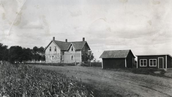 View from across road of a farmhouse and two outbuildings on an IH demonstration farm at Oak Lake. A dirt road runs past the farm and a cornfield is in the foreground.
