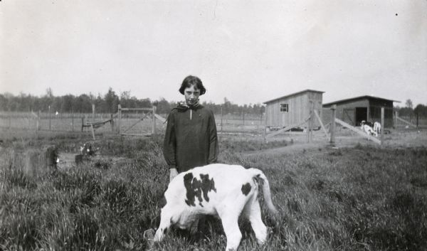 Opal Gillespie standing in a field while feeding a Holstein calf from a metal bucket. Several farm buildings and a fence are in the background.