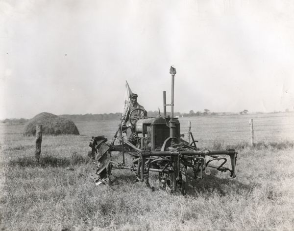 Man driving a Farmall Regular tractor with an attached cultivator to work in a field at an International Harvester demonstration farm. A fence and haystack are in the background.