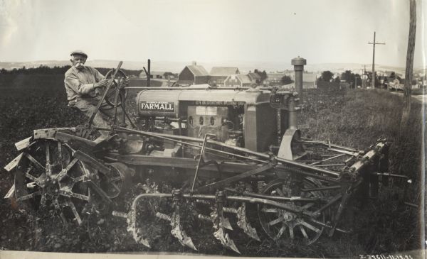 Man operating a Farmall Regular tractor with attached cultivator in a field. Decals are on the tractor. In the background are many farm buildings.