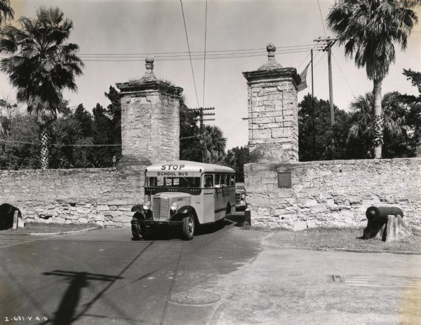 An International Model C-30 school bus passing through gates in a stone wall. Cannons stand on either side of the gate opening, and there is a plaque about the gates on the right side of the gate.