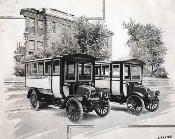 Two International Model F (or 31) buses parked side by side in front of a brick building, possibly a school. The buses are marked "Ravinia Consolidated School Dist. No.1."
