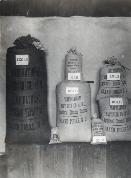 Burlap bags marked with their weight sit on a shelf awaiting shipment. The text on the bag reads: "International Harvester Co. of N.J. Agricultural Extension Department. Grand Forks, N.D."