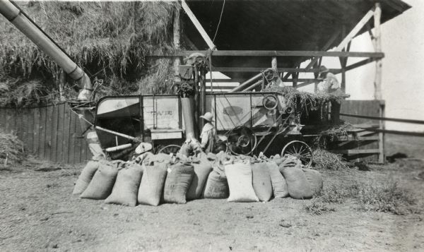 Men on an International Harvester demonstration farm are feeding corn into a husker-shredder as a boy is holding a burlap bag to the machine to catch the kernels. Bags full of corn are resting on the ground in front of the machine, and a barn is in the background.