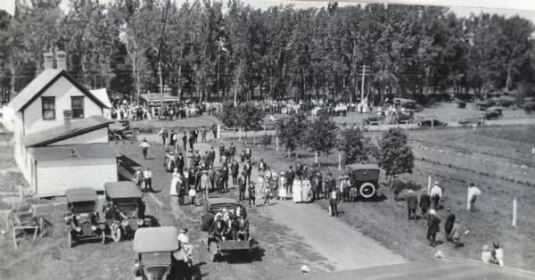 Elevated view of a crowd gathered for Picnic Day at an International Harvester Company demonstration farm. Automobiles are parked in the foreground and along the treeline in the background.