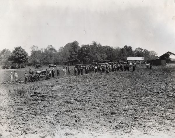 View across field on an International Harvester Company farm towards a group of men standing and watching a plowing demonstration. Several barns and other farm structures are in the background.