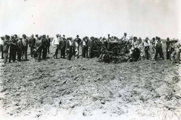 A group of men, women, and children are standing in a field at an International Harvester Company farm to watch a 4-row corn planter demonstration.