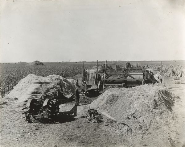 Men are using a Titan 10-20 tractor and a large piece of farm machinery, probably a thresher, to harvest a field crop on an International Harvester demonstration farm. Piles of the crop are lying in the foreground, and stacks of what appears to be hay are behind the machinery.