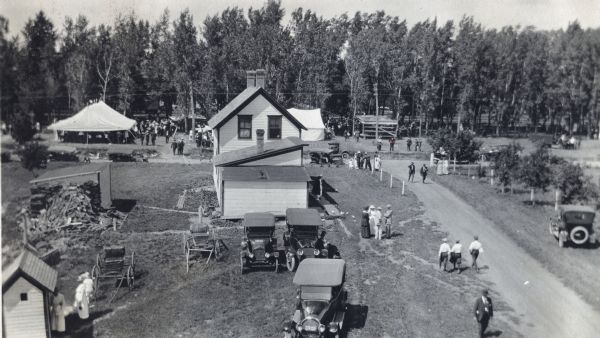Elevated view of picnic day on an International Harvester Company demonstration farm. Automobiles are parked near a farmhouse and a group is gathered near a tent in the yard and smaller buildings stand nearby.