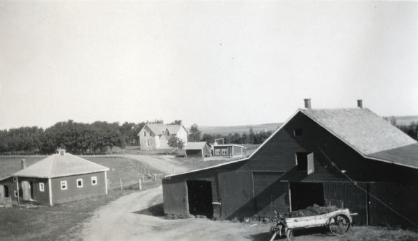 Elevated view of International Harvester Company demonstration farm. A dirt road leads to the farmhouse in the background. In the foreground, a wagon loaded with hay is parked in front of a barn and several smaller buildings stand on the property.