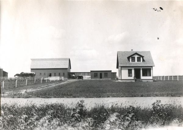 View of an International Harvester Company demonstration farm from across a dirt road. A farmhouse is to the right, and a barn and smaller buildings are nearby. An automobile is parked in the driveway leading to the buildings, and behind a fence are horses and cows. A person is sitting on the steps in the front of the farmhouse.