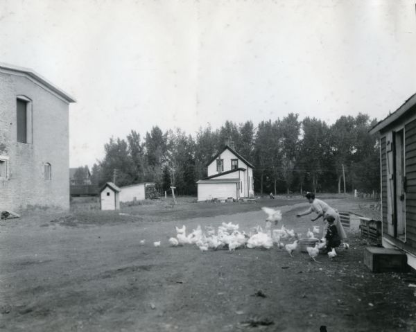 A woman and child feed a flock of chickens from a bucket while standing outside a shed. A young boy sits near the farmhouse, and a train on railroad tracks runs along the treeline in the background.