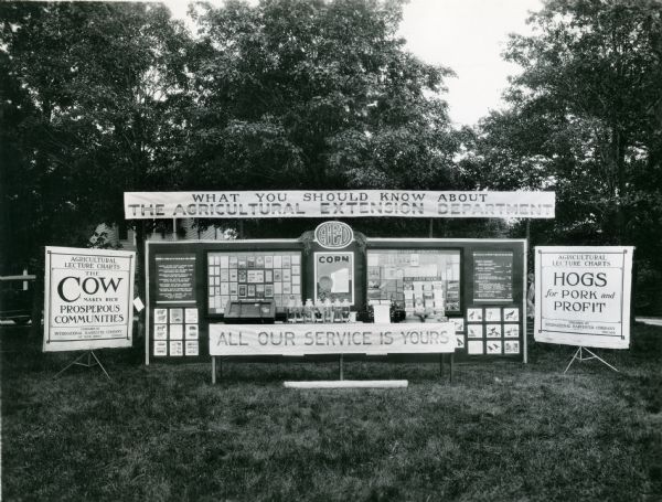 View of an Agricultural Extension Department display outdoors. Signage is displayed behind a table which appears to hold jars of samples of corn.