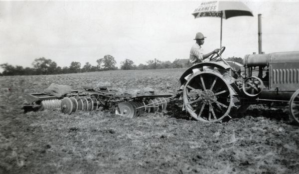 An umbrella is shading a man while he is driving a McCormick-Deering 10-20(?)tractor and disc harrow through a field.