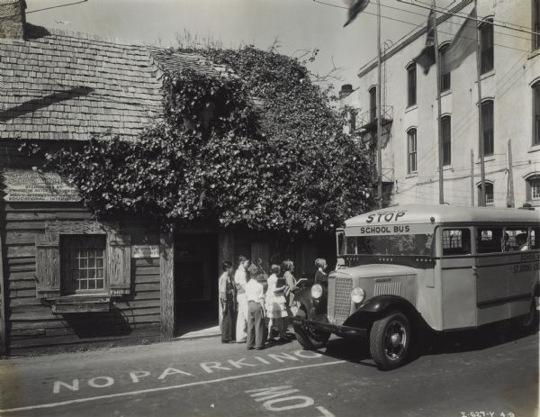 Schoolchildren boarding an International C-30 school bus. Original caption reads: "One of six Model C-30 school buses with all-steel bodies, owned by St. Johns County School Board, St. Augustive. Photograph taken in front of the oldest wooden schoolhouse in America."