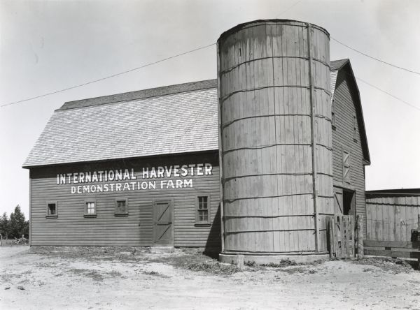 Side view of an International Harvester demonstration farm barn. A silo stands next to the barn and an additional building is in the background. The words: "International Harvester Demonstration Farm" are painted on the barn.