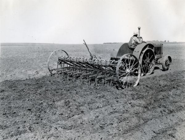 A man wearing overalls and a straw hat is pulling a field cultivator through a field with a McCormick-Deering 15-30 tractor on an International Harvester demonstration farm.