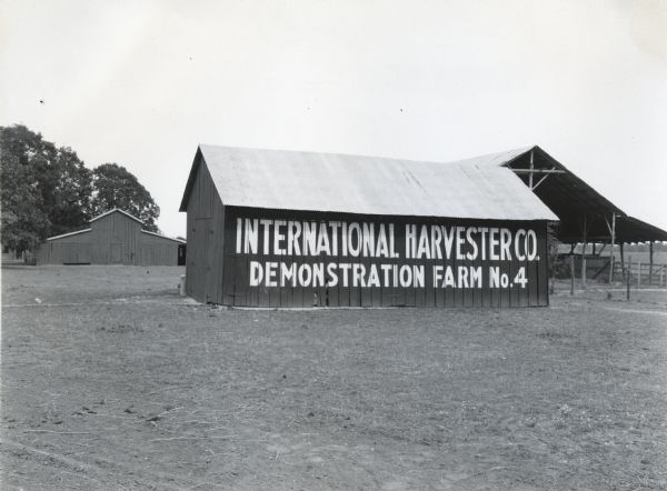 Several farm buildings stand on an International Harvester demonstration farm. The side of a wooden barn is painted with a sign that reads: "International Harvester Co. Demonstration Farm No.4."