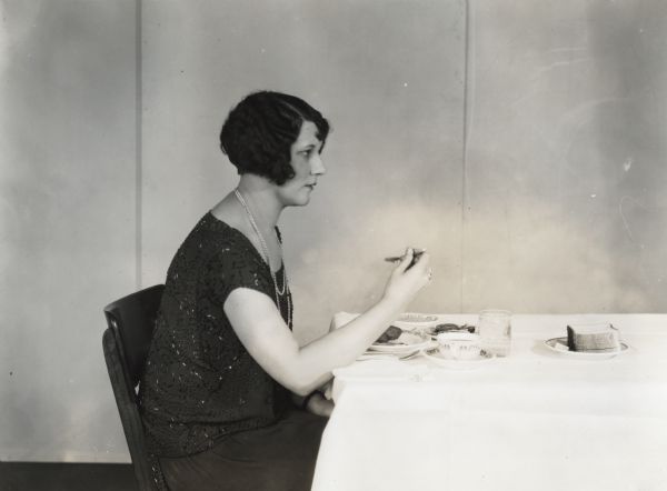 A woman wearing a beaded dress and necklace is sitting at a table to eat canned corn and peaches.
