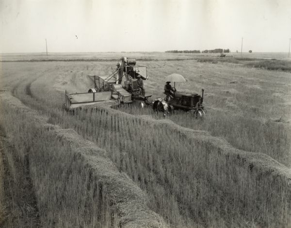 Elevated view of two men on a McCormick-Deering harvester-thresher (combine) with pickup attachment in a field. Decals and/or stencils are on the machine, including one that reads: "Always Stop Engine Before Working on or Near Moving Parts." The combine is pulled by a Farmall Regular tractor.