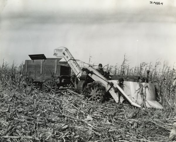 Man pulling McCormick-Deering one-row corn picker with a farm wagon in a cornfield.